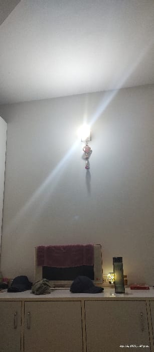 The image shows the interior of a room in Zirakpur, India (latitude: 30.629, longitude: 76.835). The room is sparsely furnished and has white walls. There is a single light fixture on the wall, which is emitting a bright light. The light is casting a long shadow across the room.  There is a TV on a cabinet and a small lamp to the right of the TV. On the cabinet are 3 caps: one black, one green, and one blue. To the right of the lamp is a black bottle and what appears to be a red book. The room is uncluttered and appears to be clean. The camera is pointed at the front of the room and is slightly angled upwards.
