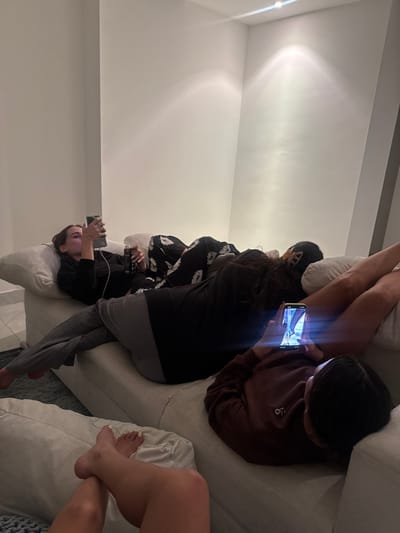 Four people lounge on a white sectional sofa in a brightly lit room. All are engaged with cell phones, their faces illuminated by the screens. One person is reclining, while the others are seated. The photo captures a casual, relaxed atmosphere.