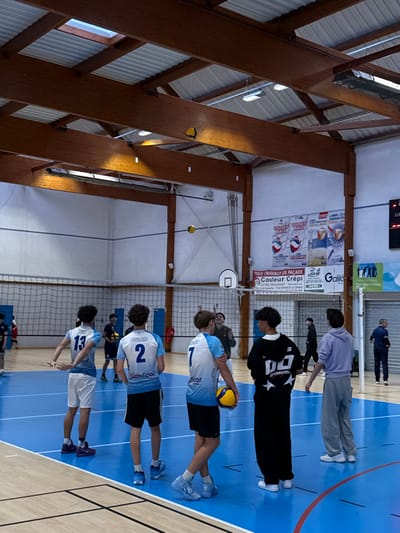 The image depicts an indoor gymnasium, specifically a volleyball court, located in Sennecey-le-Grand, France.
The scene features a blue polished sports floor with white and lighter blue court markings. A volleyball net spans the width of the court in the midground. Above the net, a basketball hoop and backboard are mounted on the far white wall. The ceiling consists of exposed wooden beams, corrugated metal panels, and skylights, with various light fixtures. A yellow and blue volleyball is lodged on one of the ceiling beams. Another yellow and blue volleyball is suspended in mid-air above the net.
Approximately ten individuals are visible. In the foreground, facing away from the camera, are five male individuals, likely players. From left to right:
1. A male in a light blue and white jersey with "13" on the back and white shorts. His arms are slightly bent at the elbows, hands held near his waist.
2. A male in a light blue and white jersey with "BOULAND" and "2" on the back and black shorts.
3. A male in a light blue and white jersey with "7" on the back and black shorts. He holds a yellow and blue volleyball in his left hand.
4. A male in a black long-sleeved shirt with white graphics ("DO EHMERS" and stars) on the back and black baggy pants.
5. A male in a light purple hooded sweatshirt and light grey sweatpants.
In the background, beyond the volleyball net, are additional individuals. To the far left, a person in dark attire is partially visible. Centered behind the net, a man with a beard, wearing a green long-sleeved shirt, observes the court with his hands slightly raised. To the far right, a man in a dark blue shirt stands near a partially visible scoreboard, which displays "LO" and "17". Another individual in dark clothing walks across the background from left to right.
The far wall features several banners, predominantly advertising "VOLLEY BALL" and "LABEL CLUB FORMATEUR PERFORMANCE," alongside an advertisement for "TOUS TRAVAUX DE FAÇADE Couleur Crépi" and "Galilé." A red fire extinguisher is mounted on the wall to the left of the net.