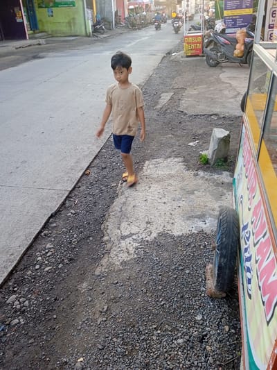 A young boy wearing a tan t-shirt, blue shorts, and yellow sandals is walking along a gravelly roadside next to a paved road. The road is in the city of Pilang, Indonesia. To the right of the boy, a yellow food cart is parked, its wheel resting on a brick. The cart has writing on its side. In the background, there are parked motorcycles and scooters, along with a sign advertising "Dimsum Mentai." Other buildings and people are visible in the distance.