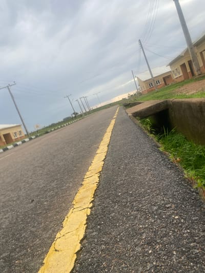 This image depicts an asphalt road with a yellow line marking its edge, stretching into the distance. The road is bordered by a concrete drainage channel on the left, filled with green grass and vegetation. Beyond the channel, several residential buildings with light-colored walls and brown accents are visible. Utility poles with power lines are also scattered along the left side of the road, suggesting an urban or suburban setting.
The perspective is low, angled towards the ground and the horizon, giving a sense of looking up from street level. The sky is overcast and appears to be either early morning or late afternoon, judging by the diffused light. The overall mood is somewhat subdued due to the cloudy sky.
The location is identified as Bulamare, Nigeria. The presence of residential buildings, paved roads, and utility infrastructure suggests a developed area within Nigeria. No people are visible in the image, and no specific activity or event is clearly taking place; it appears to be a static scene of a road and its surroundings. The image is rich in detail regarding textures and surfaces, with the rough asphalt, the smooth concrete, and the organic textures of the grass all being distinctly visible.