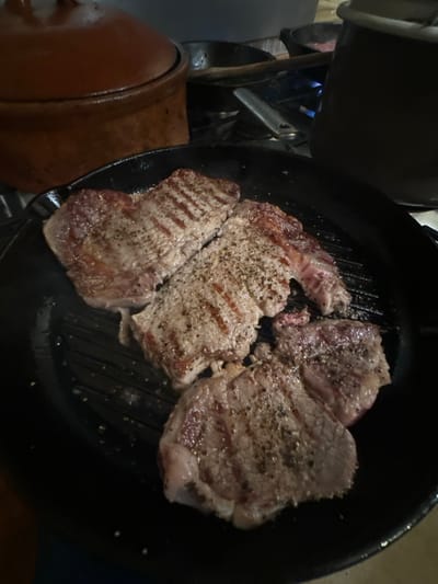 The scene depicts an indoor kitchen environment in Kfar Vitkin, Israel. In the foreground, four pieces of meat, consistent with steaks, are cooking in a black cast-iron grill pan. The meat displays visible grill marks and is seasoned with coarse black pepper. To the left of the grill pan, a large, lidded, brown earthenware pot is present. In the background, a gas stovetop is visible, with multiple dark-colored pots and pans. A distinct blue flame from a gas burner is active beneath a tall, dark pot on the right side of the background. No individuals are present in the image.