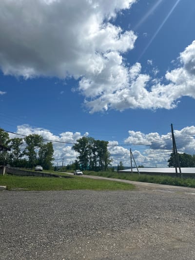 The image depicts an outdoor scene on a sunny day with fluffy white clouds against a clear blue sky. The perspective is from the side of a gravel road, looking towards a line of trees and a body of water. There are two white vehicles visible on the left side of the road, one further down the road than the other. Overhead power lines run across the sky, supported by utility poles. The surrounding vegetation is green and lush, suggesting a warm season. The lighting indicates it is daytime, likely late morning or early afternoon, due to the bright sun and strong shadows. The overall impression is of a rural or semi-rural setting, possibly near a lake or river. There is no visible text in the image.