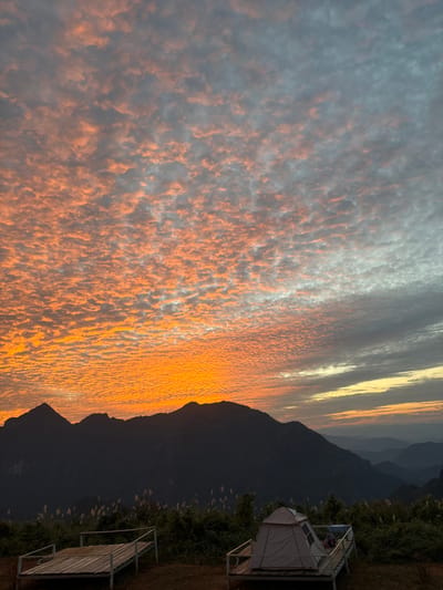 A captivating sunset displays a myriad of orange clouds against a dusky sky. Mountains form a dark silhouette along the horizon, with a tent and simple wooden structure perched on the nearby grassy terrain. The scene combines the beauty of nature with the tranquility of a campsite.
