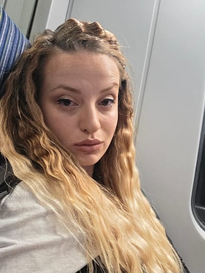 The scene depicts an individual inside what appears to be a public transport vehicle in the city of Dolni Rakovets, Bulgaria. A female subject, with fair skin and long, wavy, light brown to blonde hair, occupies the majority of the frame. Her hair is styled with a partial updo on the crown, and the remainder flows over her shoulders and chest. She has a neutral facial expression, with her dark eyes looking directly forward towards the camera. Eyeliner is visible around her eyes, and her lips are painted with a light, neutral tone. She is wearing a light-colored (likely white or off-white) garment on her left side, partially covered by what appears to be a black garment or strap on her right. To the left, a portion of a blue, white, and green striped fabric cushion, likely a headrest, is visible. To the right, a plain, light-grey or white interior wall panel with a vertical seam is present, with a dark, vertical rectangular object, possibly a window frame, partially visible on the far right. The lighting is even and consistent with an indoor setting.