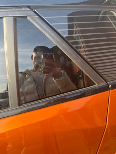The image captures a selfie reflection of a male individual in the tinted side window of a metallic orange vehicle, located in Bretton, United Kingdom. The male, exhibiting short dark hair, is wearing a light-colored puffy jacket over a t-shirt. He holds a smartphone, encased in red, in his right hand, positioned to capture his reflection. His face is partially obscured by the phone. The vehicle's window frame and B-pillar are dark. Reflected in the upper right portion of the window is a red brick building featuring multiple horizontal white slats or blinds. The upper left of the reflection shows a clear blue sky with minimal cloud cover. Faint reflections of distant vehicles or structures are discernible in the far left of the window. The scene is illuminated by direct sunlight, highlighting the car's paint and the reflections.