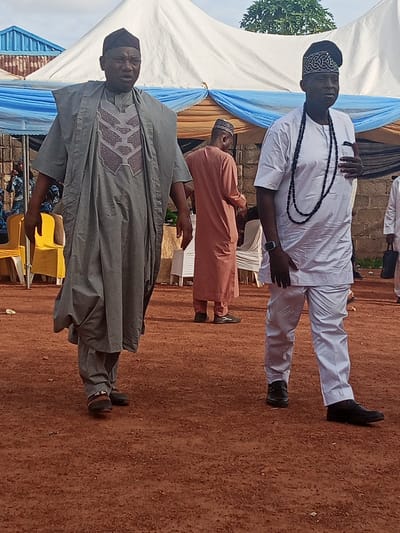 The scene is an outdoor event in Afaka, Nigeria, set on reddish-brown laterite soil. Two adult males are prominently featured in the foreground, walking from left to right. The man on the left wears a dark cap and a gray traditional agbada over a patterned tunic featuring dark brown and gray geometric designs. He is wearing dark sandals. The man on the right wears a black, embroidered cap, a white traditional Nigerian tunic and trousers, a long black beaded necklace, and a dark digital wristwatch on his left wrist. He is wearing black dress shoes, with his left hand slightly raised in a relaxed posture. In the mid-ground, a third male in a light peach-colored traditional garment is partially visible between the two main subjects. The background comprises several large white event tents, adorned with blue and gold fabric drapes. Yellow event chairs are visible under a tent to the left, and a segment of a blue-roofed building and green foliage are present further in the distance. The ground is dry, unpaved earth with minimal scattered debris.