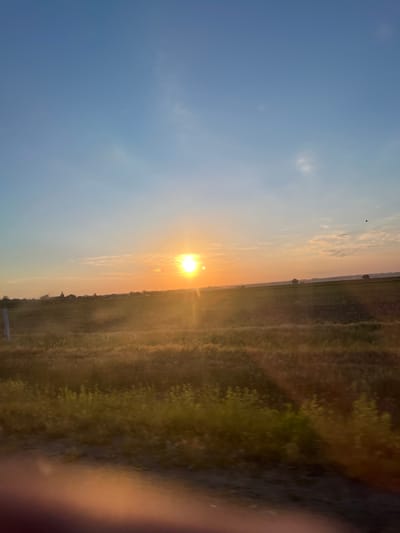 The image depicts a wide, flat landscape at dusk or dawn, likely in Karagash, Moldova. A bright sun, appearing as a stark white disk with a golden halo, is positioned in the middle ground, slightly to the right of the frame. The sky transitions from a pale blue at the top to a soft orange and yellow near the horizon, with a few wispy clouds present. A silhouette of a small bird is visible in the upper right portion of the sky.
The foreground is dominated by a field of tall, dry grass or crops, which is bathed in the warm, golden light of the setting or rising sun, casting long shadows. The texture of this field appears somewhat blurred, suggesting movement. Beyond this field, a darker, tilled field or fallow land is visible, stretching towards the horizon. Along the distant horizon line, a faint line of trees and scattered structures, possibly houses or farm buildings, can be discerned. A tall, thin vertical object, possibly a fence post or marker, stands to the left of the frame in the middle ground. The bottom edge of the image shows a blur of reddish-orange light, likely from a reflection or an object within the frame not clearly visible. The overall impression is one of rural tranquility and the natural beauty of a cultivated landscape during a transitional time of day.
