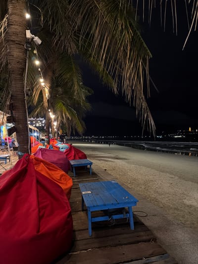 The image depicts a nighttime beach scene in Sơn Trà Ward, Vietnam, from a slightly elevated perspective on a wooden platform. In the immediate foreground and left, several large, brightly colored beanbag chairs are arranged, predominantly red, orange, magenta, and purple. Two small, weathered blue wooden tables sit directly in front of the beanbags on the wooden deck. Overhead, multiple palm trees are illuminated by strings of warm-toned bulb lights. A white security camera and an additional light fixture are mounted on the trunk of the nearest palm tree. Further down the illuminated line of trees and beanbags to the left, indistinct figures of individuals are visible, some seated, under a temporary tent structure.

The midground features a wide, unpopulated sandy beach extending to the right and into the distance. Gentle ocean waves are visible at the water's edge, reflecting faint light. One or two distant, silhouetted figures can be observed walking on the sand. The background consists of a dark night sky and a distant coastline marked by numerous scattered city lights. A distinct, brightly lit structure, possibly a monument or large building, is prominent on a elevated landmass to the far right.