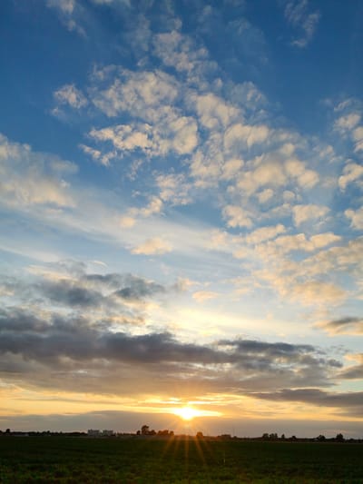 The image captures a sunset over a field near the city of Pinsk, Belarus. The sky is filled with a dynamic mix of blue, white, and golden clouds, with the sun peeking through on the right side, casting long rays. The foreground consists of a dark green field. In the distance, a low-lying horizon shows a few trees and buildings, hinting at a rural or suburban landscape. The overall atmosphere is serene and natural.