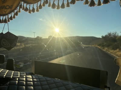 The image is a forward-facing view from inside a vehicle, likely a truck, looking out the windshield. The sun is setting or rising directly ahead, creating a strong glare and lens flare that obscures details of the distant landscape and sky. The sky is clear and blue. In the foreground, the quilted interior of the vehicle is visible, with dark upholstery. Hanging from the top of the windshield is a decorative trim with tassels and a logo that appears to read "SCAN" with a crown. Two dice-shaped air fresheners are also hanging from the windshield. The windshield itself is visibly dirty, with streaks and smudges. The road ahead is paved and curves to the right, with dry, grassy hills and sparse vegetation on either side. A utility pole is visible in the middle distance. The image appears to have been taken on a road in or near Avra, Greece, based on the context provided.
