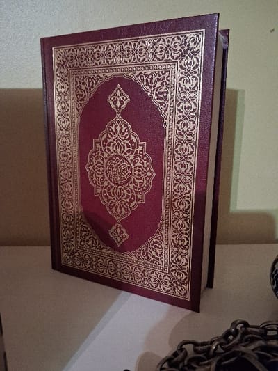 A close-up shows a burgundy book with gold ornate designs. The book cover features a large oval center with intricate floral patterns and calligraphy. The borders are also decorated with gold geometric and floral designs. The book appears to be thick and heavy. Part of a metal chain is visible in the background. The background is primarily a wall, casting soft shadows.