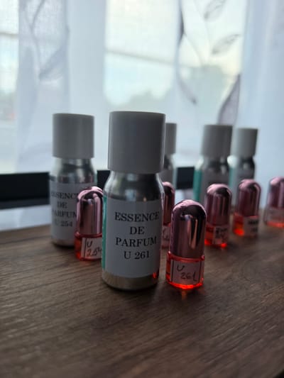 The image shows a collection of small glass perfume bottles with metallic pink caps arranged on a wooden surface. Some of the bottles have labels with text and numbers. In the background, a sheer white curtain is visible, with diffused light coming through it. The scene appears to be indoors, likely a well-lit room. There are no people or animals visible in the image. The overall impression is one of organized display or storage of fragrance samples.
