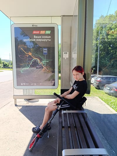 A young female with dyed red hair sits on a metal bench at a bus stop. She is wearing a black t-shirt and black shorts with white stripes. A black scooter is propped up between her legs. In the background, a large digital display shows a map of new bus routes. The bus stop has a glass enclosure and is surrounded by trees and parked cars. The ground is paved with concrete and asphalt. The sky is clear and blue. The scene appears to be in Shishkin Les, Russia, as indicated by the map on the bus stop display.