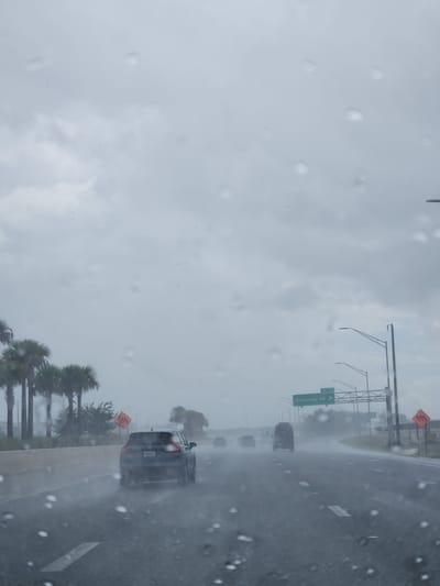 The image depicts a highway scene in Orlando, Florida, during a rainstorm. The perspective is from inside a vehicle, with raindrops visible on the windshield, creating a blurred effect. Cars are driving on the wet asphalt highway under a cloudy, overcast sky. Palm trees are visible along the side of the road. Highway signs are present, one of which indicates "Exit 12 Goldenrod Rd". Construction barrels and signs are also visible along the roadside, suggesting ongoing work. The visibility is reduced due to the rain, creating a hazy atmosphere.