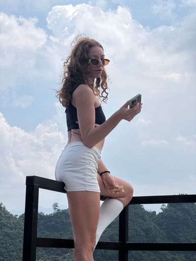A woman wearing a black crop top, white shorts, and tall white socks sits on a black metal structure in Lũng Phìn Commune, Vietnam. She is holding a smartphone in her left hand and a cone of some kind in her right. The background consists of a partly cloudy sky and a view of a mountain through the metal structure. The image is oriented sideways.