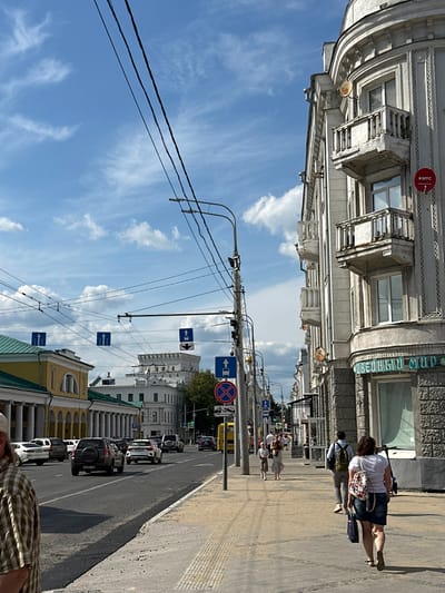 This image depicts a street scene in Golovinskoe, Russia, under a partially cloudy sky. A wide, paved road runs through the center of the frame, with several cars driving in both directions. The sidewalk on the right side of the road is populated by pedestrians walking in various directions.
On the right side of the street, a light-colored, multi-story building with ornate architectural details and balconies dominates the foreground. A red circular sign with white lettering and a number is attached to the building. Further down the street, another similar building is visible.
On the left side of the street, a row of buildings with a yellow facade and a green roof features arched windows and columns. Beyond this, more buildings are visible, including a prominent white structure with a tower-like element.
Numerous power lines and traffic signals are suspended across the street, indicating an urban environment. Traffic signs, including directional arrows and a prohibition sign, are also present. A yellow bus is parked on the side of the road. The overall impression is a bustling city street on a bright, sunny day.