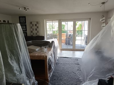 A room in Essenbach, Germany, is prepared for painting or renovation. Furniture, including a table and a sofa, is covered with clear plastic sheeting for protection. A dark gray protective sheet covers the floor. Natural light floods the room through glass doors that lead to a balcony. Decorative wall art is visible on the left wall.