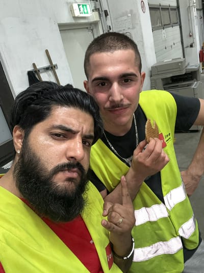 Two men are posing for a selfie in what appears to be a warehouse or industrial setting in Schweitenkirchen, Germany. The man in the foreground on the left has a full beard and dark hair, wearing a red shirt and a high-visibility yellow vest. The man on the right has short dark hair and a mustache, also wearing a high-visibility yellow vest over a black shirt. He is holding a stroopwafel in his right hand, bringing it towards his mouth. The background shows industrial doors, a copier or printer, and an emergency exit sign. The lighting is artificial, suggesting an indoor environment.