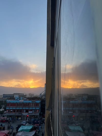 This is a high-angle, vertical-format photograph of a city street scene at dusk, viewed through a window with a reflection of the same scene. The location is the city of Kabul, Afghanistan. The sky is a gradient of pale blue to orange and yellow as the sun sets behind a mountain range in the distance. Dark, textured clouds are visible in the sky, illuminated from behind by the setting sun.

In the foreground and midground, a busy street market is depicted. Buildings with a blue facade and red accents are prominent, with multiple levels of windows. Some buildings have signage with Arabic script. Numerous people are walking and interacting in the market area. Several stalls are set up, covered with colorful tarps, selling various goods. Vehicles, including cars and vans, are present on the street, some of which appear to be taxis or delivery vans. The scene conveys a sense of daily life and commerce in an urban setting.

The right side of the image features a large, reflective window, showing a distorted reflection of the street scene and the sunset. The reflection is slightly hazy and captures the same elements as the actual scene, but with a different perspective. A portion of a building frame, which appears to be made of wood and concrete, is visible on the far right and is also reflected in the window. The lighting suggests late afternoon or early evening.