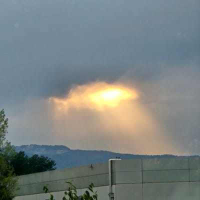 The image captures a sky with dramatic lighting, likely due to clouds. A bright, sunlit area pierces through a dense cloud formation, creating a beam of light. In the foreground, a building with a light-colored facade is visible, possibly a commercial or industrial structure. The building has a segmented design with visible lines between sections. To the left of the building, green foliage, including trees and smaller plants, is present, suggesting an outdoor setting within or near a populated area. In the background, a mountain range is silhouetted against the sky. The overall impression is a natural landscape juxtaposed with urban infrastructure. The specific location in Vic, Spain, is not discernible from the image itself but is provided as context for the scene.