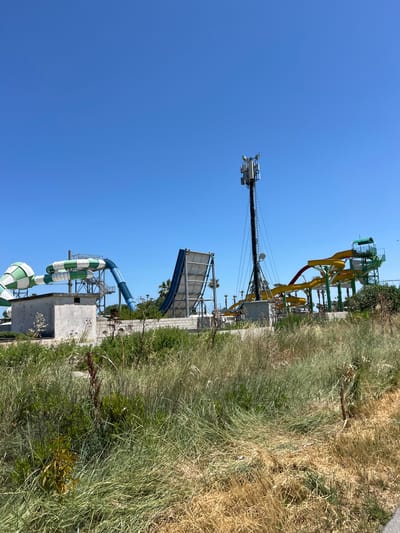 This media file captures a sunny day at what appears to be a water park in Manfredonia, Italy. The primary subjects are the water slides and the surrounding environment.

Several water slides are visible, including bright yellow and red tubular slides at the top of the frame, and a blue slide lower down. There are also green and white striped tube slides in the foreground. These slides are all elevated on a metal frame structure.

In the background, a tall communication tower with antennas is prominent against a clear, bright blue sky. The weather appears to be fair and sunny, suggesting it's likely daytime, possibly late morning or early afternoon, given the intensity of the light and the deep blue of the sky.

The scene is bordered by dry, tall grasses and some greenery, suggesting a somewhat arid or natural landscape adjacent to the constructed water park facilities. The presence of palm trees in the background further supports a warm climate location. There is no visible text in the image. The overall impression is one of a recreational facility on a pleasant, sunny day.