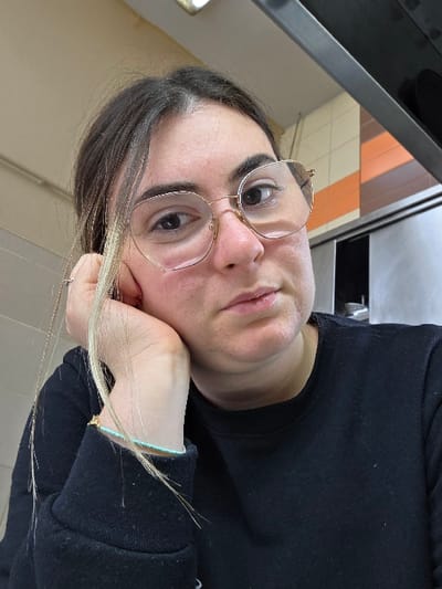 A female subject is depicted in a close-up, chest-up portrait, located in a commercial or back-of-house indoor setting in the city of Gevgelija, North Macedonia. The subject has dark brown hair with visible lighter strands or streaks framing her face and is wearing round, gold-rimmed eyeglasses. She is dressed in a black long-sleeved sweatshirt. Her right hand is positioned with her palm supporting her chin, and a light blue beaded bracelet with a small gold-colored charm is visible on her right wrist. The background features wall tiling consisting of light beige squares and a section of horizontal orange and brown stripes. Above the tiled wall, a metallic structure, consistent with a commercial kitchen hood or shelving, is present. A fluorescent light fixture is partially visible on the ceiling in the upper left quadrant. The subject's gaze is directed towards the camera.