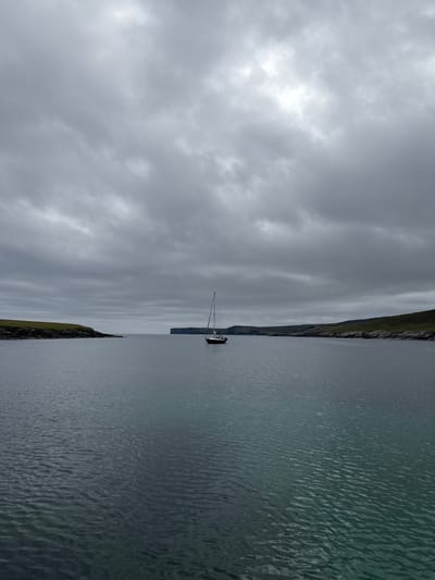 This media file captures a tranquil scene of a sailboat on a body of water, likely the sea, given the location context of Shetland, United Kingdom. The sailboat, a solitary object, is the main focus against the backdrop of a dramatic sky and a rugged coastline. The sky is filled with heavy, overcast clouds, suggesting a dull or possibly stormy weather condition. The water is calm, with subtle ripples on its surface, reflecting the muted light from the sky. A steep, green-covered cliff face runs along the edge of the water, indicating a coastal landscape. The overall mood is one of quiet solitude and the raw beauty of nature. There is no visible text in the image. The time of day is difficult to ascertain precisely due to the overcast conditions, but it appears to be daytime.