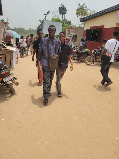 The image depicts an unpaved, light brown dirt road in Candelaria, Colombia, under bright, hazy daylight. The scene features multiple individuals and motorcycles, suggesting a bustling outdoor environment.

In the foreground, two young adult males walk towards the camera. The male on the left, of dark complexion, wears a light blue plaid short-sleeved button-up shirt, dark patterned trousers, and dark open-toed sandals. He carries a dark brown messenger bag with a repeating monogram pattern across his right shoulder. He looks directly at the camera. To his right and slightly behind him, a second young adult male, also of dark complexion, wears a dark short-sleeved t-shirt, dark trousers, and open-toed sandals. He has a small dark crossbody bag with a purple strap on his left side.

In the mid-ground, to the left of the foreground subjects, another young adult male in a dark t-shirt and light shorts walks. Further back on the left, a person on a light-colored motorcycle is partially visible, holding a light blue umbrella for shade. The rear of a dark motorcycle with a partially visible license plate (e.g., "K 11704") is parked in the far left foreground.

On the right side of the image, a building with a red lower section and a lighter upper section features two dark, rectangular openings, possibly shuttered windows. Above these openings, a sign is partially visible. Several motorcycles are parked or being ridden near this building. A young adult male, wearing a white t-shirt and dark trousers, with a multicolored crossbody bag, walks away from the camera towards the right side of the frame, near the building.

The background includes other structures, dense green vegetation, and tall palm trees against a light, hazy sky. Various unreadable signs and posters are visible on walls and posts throughout the scene. Shadows of people and objects are cast on the dirt road, indicating sunlight from the upper right.