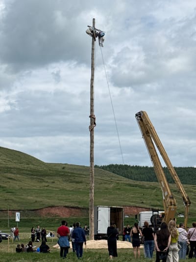 This image depicts an outdoor event in Nurykuevo, Russia, under a cloudy sky. The central focus is a tall wooden pole, at the top of which two individuals are positioned. One person is actively climbing the pole, clinging to it with their arms and legs. Another person is near the top, holding onto a cross-like structure. A rope extends from the top of the pole downwards.
In the foreground, a crowd of spectators watches the event. People are scattered across a grassy field, some seated, others standing. Several individuals are dressed casually, indicating a relaxed, community gathering.
Behind the crowd, a large yellow crane is visible, its arms extended. Nearby, a white truck and a smaller white vehicle are present, suggesting logistical support for the event. The background features rolling green hills and a dense line of trees. The overall scene suggests a local festival or competition involving pole climbing.