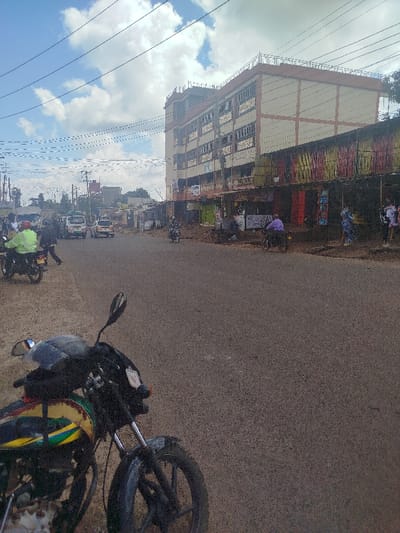 This image captures a street scene in Kihara ward, Kenya, on a partly cloudy day. The foreground features the front section of a parked motorcycle on the left, displaying a yellow, green, and black fuel tank. The asphalt road occupies the central and right portions of the lower frame.

Along the left side of the road, multiple individuals are visible. One person in a neon green jacket is mounted on a motorcycle. Further down, several white vans, likely public transport vehicles, are present on the road. Utility poles with numerous overhead wires extend across the upper left and central sky.

The right side of the street is dominated by a multi-story building, approximately five floors tall, with a light-colored facade and brown horizontal banding. Below this structure, a row of single-story commercial establishments or stalls with corrugated metal roofs, some painted in vibrant stripes of red, yellow, and green, line the thoroughfare. Individuals are visible near these shops; one person in a purple shirt rides a bicycle, while others stand. Another motorcycle is observed further down the street on the right. The ground immediately adjacent to the shops appears unpaved. The background shows additional buildings and trees under a partly cloudy sky, with the road receding into the distance.