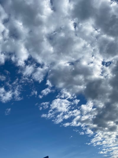 The image shows a bright blue sky filled with a mix of white and gray clouds. Some clouds are thin and wispy, scattered across the upper part of the frame, while others are thicker and more dense, appearing on the right side of the image. The lower portion of the sky is clear blue. A small portion of a dark blue sign with a white edge is visible at the very bottom of the image. The scene appears to be an outdoor view, likely from ground level, looking upwards. The context provided places this scene in the city of Dobieżyn, Poland.