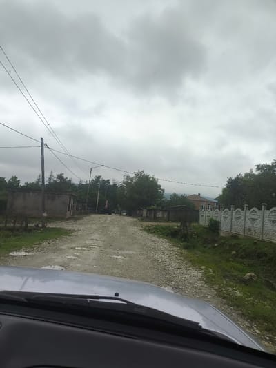 A paved road stretches through a village under a cloudy sky. The road surface is uneven, with loose gravel and some puddles indicating recent rain. On the left side of the road, a weathered, rectangular building with a flat roof sits partially overgrown with greenery. Beyond this structure, a wooden fence runs along the property line, and further back, lush trees line the horizon. A wooden utility pole stands tall, adorned with electrical wires that crisscross overhead. Further down the road, a red tractor with a loader attachment is parked, and a few indistinct figures can be seen walking. On the right side of the road, a decorative concrete fence with a scalloped design borders a property. Behind the fence, trees and foliage are visible, and in the distance, a brick house with a tiled roof can be seen. The foreground of the image shows the hood and windshield of a silver car, suggesting the photo was taken from inside a vehicle. The overall impression is that of a rural, somewhat unkempt village scene on an overcast day in Skurdi, Georgia.