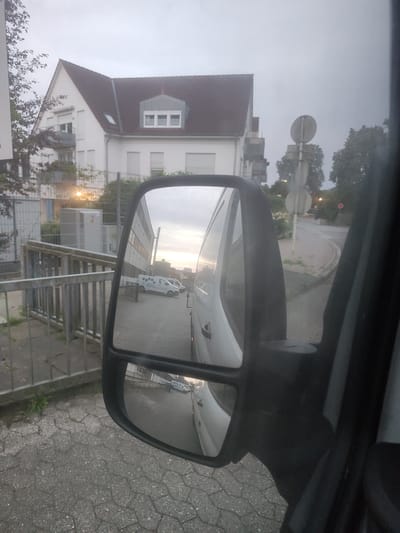 The image is a medium shot of a side-view mirror on a vehicle, reflecting a street scene in Eutin, Germany. The mirror is attached to a black vehicle and shows a reflection of a white building with a red roof, some trees, and street signs. In the reflection, there are two white vans parked on a street. One van appears to be a delivery van with a logo on its side. The street is paved with cobblestones. The sky is overcast, suggesting it may be early morning or late evening, or a cloudy day. The building in the reflection has multiple windows and balconies. The overall scene is an ordinary urban environment, likely on a street in Eutin.