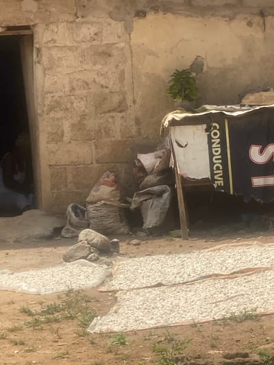 The scene depicts an outdoor area in Idiroko, Nigeria, under bright daylight conditions. In the foreground, two rectangular sections of light-colored material, possibly tarps or woven mats, lie on dry, reddish-brown soil, covered uniformly with small, irregularly shaped, light-colored pieces, consistent with agricultural produce drying. Sparse green vegetation grows on the surrounding ground.
To the left, a building with a rough, unpainted, light-brown block wall is visible. An open doorway reveals a person seated within, partially obscured, wearing light-colored attire. Against the building's wall, to the right of the doorway, several large sacks are present; one appears to be a woven sack with red and white markings, while others are grey. A small pile of grey rocks rests on the ground in front of these sacks.
Further to the right, a makeshift structure is erected, supported by a wooden pole. It features a black covering with a vertical white section displaying the word "CONDUCTIVE" in black capital letters, with a large, partially visible white "S" to its right. A lighter fabric drapes over the top of this structure. Behind or on top of this structure, against the wall, a small green plant is visible.