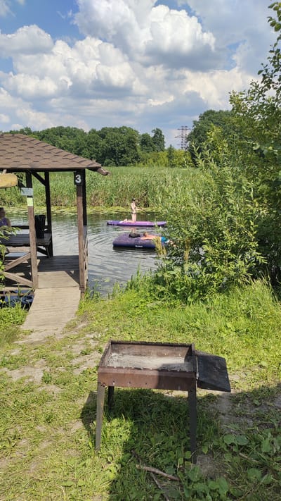 In Ramon, Russia, on a sunny day, a riverside scene unfolds. A wooden gazebo with a shingled roof stands on the left, offering shelter with a picnic table inside. A small "3" is affixed to one of its posts. Adjacent to the gazebo, a rusty metal barbecue grill sits on the grassy bank. The water of a river or lake is calm, with reeds lining the far bank. Two individuals are enjoying the water: one person is lying on an inflatable mattress, and another is kneeling on a paddleboard, holding a paddle. The sky above is blue with scattered white clouds.