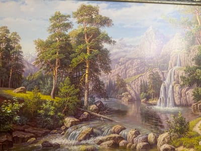 This is a highly detailed oil painting of a majestic mountain landscape with a river and waterfalls. The scene depicts a pristine natural environment, likely a wilderness area.

In the foreground, a rocky river flows over a series of small cascades, creating white foam and spray. Large, smooth boulders are scattered throughout the riverbed and along its banks. Lush green vegetation, including bushes and small trees, grows on the left side of the river. A fallen tree trunk lies partially submerged in the water.

Further back, the river widens and flows into a larger body of water. On the right bank, a dramatic waterfall cascades down a steep cliff face, feeding into the river. The cliffs are rugged and covered with scattered trees and smaller vegetation.

In the mid-ground and background, towering mountains rise, their peaks partially obscured by mist and clouds. Dense evergreen forests line the lower slopes of the mountains. Tall, mature pine trees dominate the left side of the composition, their branches spread wide. The sky is a soft blue with scattered white clouds, suggesting a clear day with some atmospheric haze.

There are no people or animals visible in the painting. The overall impression is one of serene natural beauty and untouched wilderness. There is no indication in the painting that this scene is located in the city of Svobody, Russia. The style of the landscape is generic to mountainous regions with rivers and waterfalls and could be found in many parts of the world.