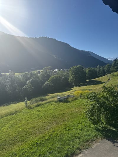 This is a landscape photograph taken in the city of Saint-Jean-d'Aulps, France. The image captures a sunlit valley with rolling green hills and a forested mountain in the background. In the foreground, a paved pathway winds its way up a grassy slope, with a group of three people walking along it. Several trees are scattered across the landscape, and in the middle ground, there are some buildings that appear to be part of a small village. The sun is shining brightly, casting rays of light across the scene. The sky is clear and blue.