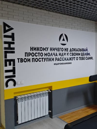 This image captures an interior wall within an athletic facility in Pyt'-Yakh, Russia. The upper portion of the wall exhibits a light-colored brick texture, transitioning to a smooth white surface, a prominent yellow horizontal stripe, and a dark gray lower section. The ceiling features a grey grid-patterned tile, and the floor comprises light-colored flooring, with a section of wooden planks visible on the right.

Prominently displayed on the white wall is a black circular logo containing a stylized "A" or delta symbol. Below this, large black Cyrillic text reads: "НИКОМУ НИЧЕГО НЕ ДОКАЗЫВАЙ. ПРОСТО МОЛЧА ИДИ К СВОИМ ЦЕЛЯМ. ТВОИ ПОСТУПКИ РАССКАЖУТ О ТЕБЕ САМИ." This translates to: "Don't prove anything to anyone. Just silently go towards your goals. Your actions will speak for themselves." Below the quote, smaller black text identifies the author as "ФЕДОР ЕМЕЛЬЯНЕНКО" (Fedor Emelianenko).

To the left, the word "ATHLETIC" is vertically oriented in large, black, three-dimensional letters. A white double light switch is mounted on the white wall section above the yellow stripe. A white, multi-fin radiator is fixed to the lower dark gray wall, directly beneath the yellow stripe, with visible plumbing connections. On the far right, a segment of a black treadmill, including a handrail, is visible, positioned on the wooden floor section. The scene is devoid of any individuals.
