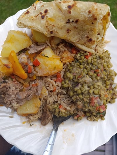 A close-up shot captures a generously portioned meal served on a white, fluted-edge plate, likely enjoyed outdoors in Kaplelwo, Kenya. The main components include a folded, golden-brown flatbread, resembling chapati, resting atop a bed of aromatic long-grain rice. This rice dish appears to be a pilau or biryani, intermingled with tender pieces of meat (possibly beef or goat), diced red and orange bell peppers, and large chunks of stewed potatoes and possibly carrots.
Alongside the rice and meat, a distinct portion of stewed green gram, locally known as ndengu, is visible, seasoned with diced tomatoes and red onions, giving it a rich, textured appearance. A silver spoon is partly submerged in the food, suggesting the meal is actively being consumed. The background shows lush green grass, reinforcing the outdoor setting. A person's arm or leg, wearing a dark blue and white patterned fabric, is subtly visible at the bottom left, holding the plate. The natural lighting indicates it's daytime. The combination of dishes strongly points to a traditional East African meal.