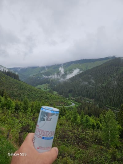 The image shows a mountain landscape on a cloudy day in Vyšná Boca, Slovakia. In the foreground, a hand holds a can of "Spring Edition" cherry soda. The background features a valley with forested slopes partially obscured by clouds. A winding road is visible through the trees. The watermark "Galaxy S23" appears in the bottom right corner of the image.
