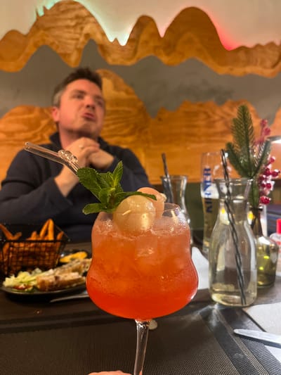 An orange-hued cocktail with ice, lychee fruit, and a sprig of mint sits in a glass with a clear straw. Behind the drink, a plate holds sweet potato fries and sandwich halves. A man in a dark blue shirt is seated to the left, with an ornamental wall behind him. A bottle with a sprig of red berries and pine needles is in the background, next to a gray tablecloth. The image is rotated clockwise.