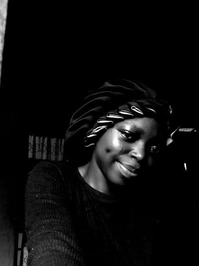 A young woman with dark skin wears a bonnet on her head. The bonnet has white trim around the edge. She is wearing a black sweater. She has a dimple on her left cheek. She has dark eyes and lips. She is smiling slightly and appears to have a ring on her tongue. The background is dark.