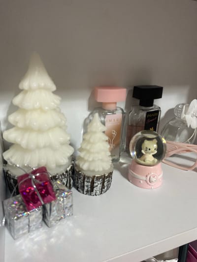The image displays a collection of items arranged vertically. In the foreground, a white, Christmas tree-shaped candle is prominently featured, with two small, shiny gift-wrapped boxes, one pink and two silver, placed beside its base. Behind this candle, another, smaller Christmas tree-shaped candle with a dark, textured base is visible. Above these candles, two perfume bottles are stacked. The upper bottle has a black cap and contains clear liquid, with the label "IMARI Cosnt" visible. It is topped with a clear spherical ornament containing a small, yellow bear figurine. Adjacent to this is a pink, rounded object with decorative white lines and a small, white figure. The lower perfume bottle has a pink cap and contains a light brown liquid, with a label that partially reads "IMARI". The background is a plain white surface, suggesting an indoor setting. The lighting is bright, indicating daytime or well-lit interior conditions. There are no people visible, and the scene appears to be a curated arrangement of decorative and cosmetic items.