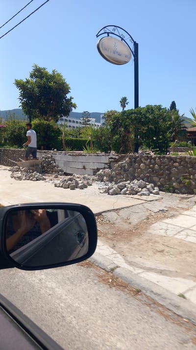 In the city of Heraklion, Greece, on a bright, sunny day, a construction site is visible by the side of a road. A man in a white t-shirt and shorts is working near a wheelbarrow and a pile of stones. Part of a stone wall is under construction, with some sections made of large stones and others of concrete blocks. Debris, including scattered rocks and broken concrete, litters the ground. In the foreground, a car's side mirror reflects a person's hands and arms, suggesting the photo was taken from inside a vehicle. A sign with text, possibly indicating a business name like "Crystal Palace," is suspended above the scene. Lush green trees and foliage surround the construction area, and a white multi-story building can be seen in the background, nestled against a hillside. The overall scene depicts ongoing renovation or construction work in a semi-urban environment.