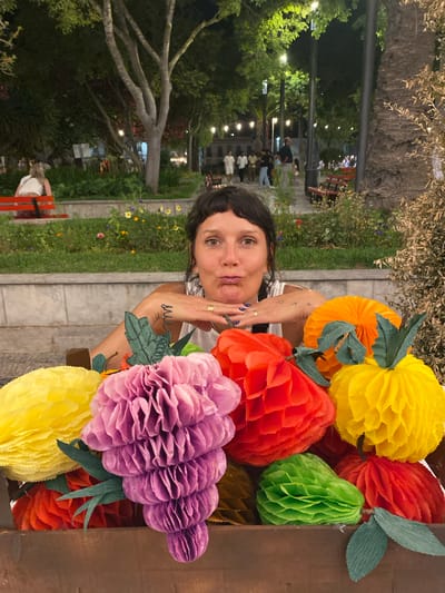 In the city of Tavira, Portugal, a woman is pictured in the foreground with a playful expression, her hands resting on her chin. She is wearing a white top and has a gold ring on her left ring finger. A tattoo is visible on her left forearm. Behind her, a wooden planter is filled with colorful, honeycomb-style decorative fruits in shades of yellow, pink, red, and green. In the background, a park setting is visible with trees, flower beds, and people seated on benches and walking. Streetlights illuminate the scene, suggesting it is evening.