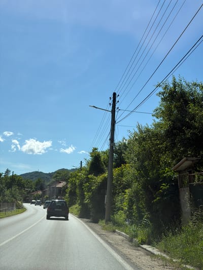 The image depicts a rural road in Beli Osam, Bulgaria, under a clear blue sky with sparse clouds. Several cars are visible driving on the road, with one black SUV in the foreground. Overhead, power lines stretch across the frame, supported by a concrete utility pole. Dense green trees line the right side of the road, with some buildings and rolling hills in the background. The scene appears to be a sunny day, likely in a semi-urban or village setting.