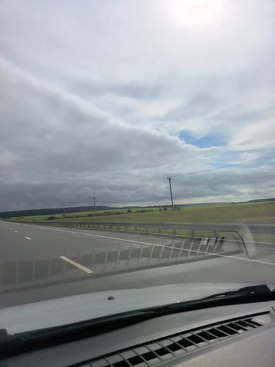 The image shows a view from inside a vehicle traveling on a highway. The road is asphalt with white lane markings. On the right side of the road, there is a metal guardrail and a green field with some trees and power lines in the distance. The sky is overcast with some patches of blue sky. The foreground shows the interior of the car, including the dashboard and windshield. The location is the M5 highway near the village of Nyestukovo in Russia.