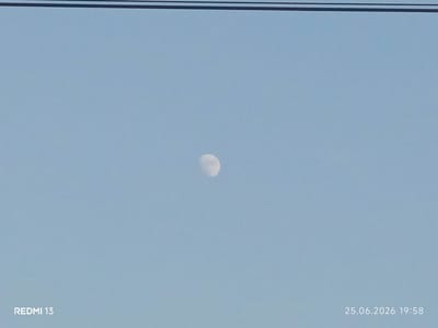 The image shows a pale, crescent moon against a clear, light blue sky. The moon is a bright, white orb with some visible craters and texture. It is positioned slightly to the left of the center of the frame, high in the sky. In the foreground, a thin, dark horizontal line runs across the top of the image, suggesting a wire or a power line.
In the bottom left corner, the text "REDMI 13" is visible in white lettering, indicating the device used to capture the image. In the bottom right corner, the date and time are displayed in white text: "25.06.2026 19:58". This suggests the photograph was taken in the early evening, as indicated by the time and the presence of the moon in the sky. The overall scene is serene and minimalist, with the sky dominating the frame and the moon as the sole celestial subject. The weather appears clear, with no clouds present. The location is identified as Belovodskoe, Kyrgyzstan.