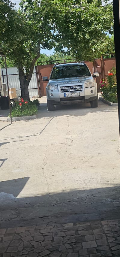 The image shows the front of a silver Land Rover Freelander parked on a paved surface. The vehicle has a license plate with the text "AX 4692 IT". The car is positioned in what appears to be a yard or driveway, with a large tree with green foliage on the left side of the frame. Behind the tree and to the right of the car, there is a reddish-brown structure, possibly a fence or building, and some pink and red flowers blooming. The ground is a mix of concrete and a mosaic-like stone pattern. The lighting suggests it is daytime, with shadows visible on the ground. The scene appears to be in a residential or semi-rural setting. There are no people visible in the image. The overall impression is a still, outdoor scene during daylight.
